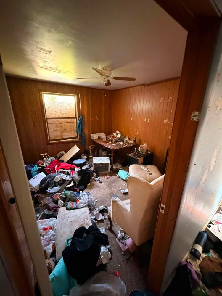 Wood-paneled back bedroom in an Elkton, Maryland cleanout, heaped with clothing, a mattress, an armchair and a window air conditioner